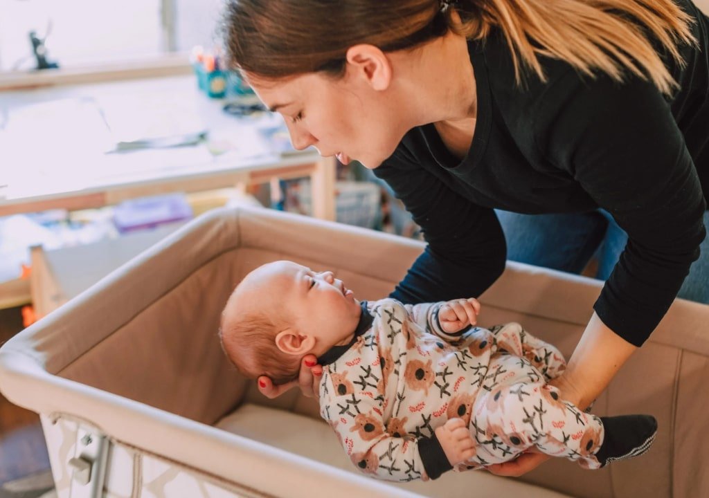 Pureday En kvinde læner sig over for at interagere med en baby i en tremmeseng, klædt i en blomstret trøje. Scenen, der legemliggør naturlig velvære, udspiller sig i et hyggeligt, godt oplyst rum.