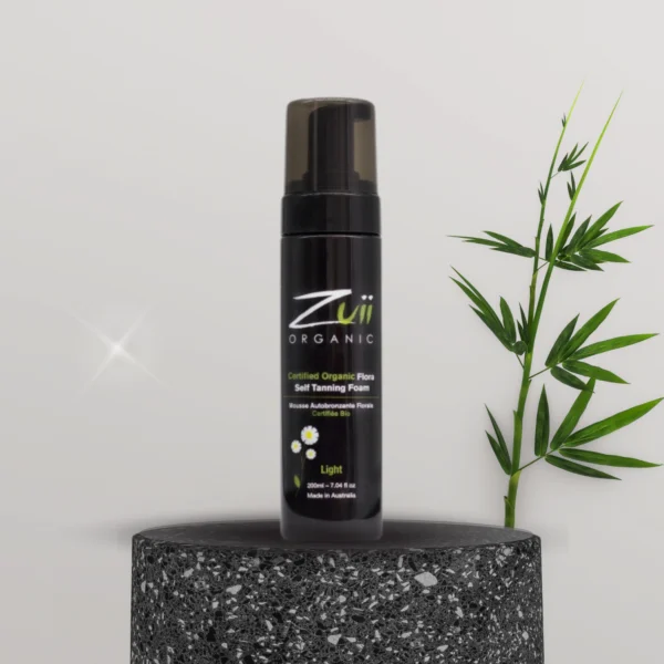 Pureday En flaske Zuii selvbruner mousse lys vist på en tekstureret grå piedestal, med en grøn bambus stilk til højre, mod en lys grå baggrund, der inkarnerer økologi og wellness.