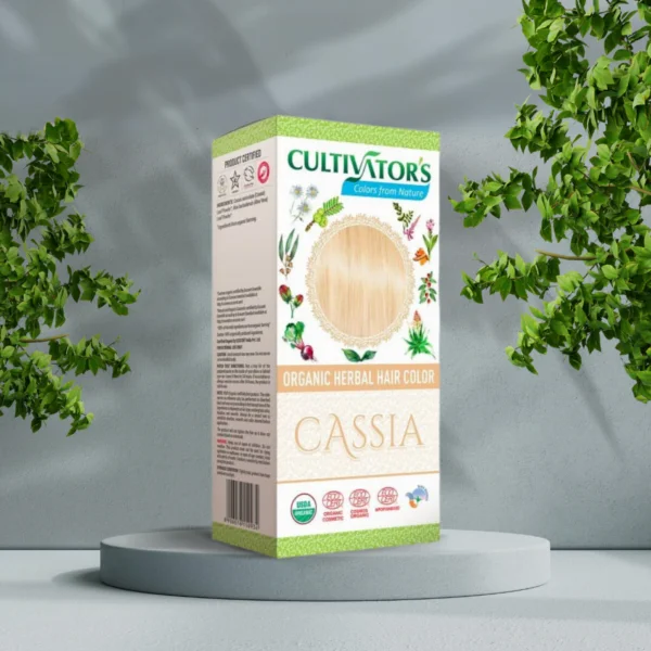 Pureday En æske med Cultivators Cassia økologisk urte-hårfarve vist på en cirkulær piedestal med en baggrund af grønne blade og en tekstureret grå væg, der understreger dens rendyrkede essens.