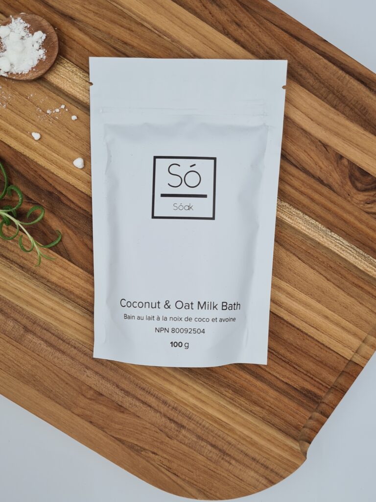 Pureday En hvid pose mærket "Coco Cream Soak 130g" er placeret på en træoverflade, der inkarnerer Pureday-oplevelsen. Frisk rosmarin og en knivspids hvidt pulver er i nærheden, hvilket forbedrer de økologiske spa-lignende omgivelser.