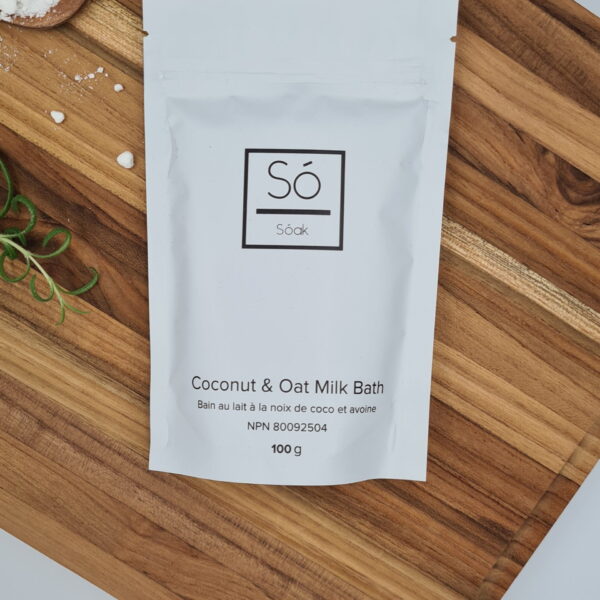 Pureday En hvid pose mærket "Coco Cream Soak 130g" er placeret på en træoverflade, der inkarnerer Pureday-oplevelsen. Frisk rosmarin og en knivspids hvidt pulver er i nærheden, hvilket forbedrer de økologiske spa-lignende omgivelser.