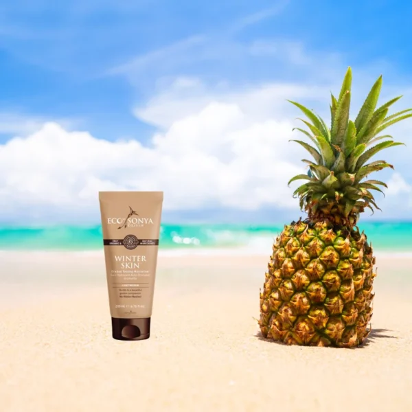 Pureday En flaske Winterskin - gradvis selvbruner lotion stående ved siden af en ananas på en sandstrand, med en klar blå himmel og turkisblåt hav i baggrunden.