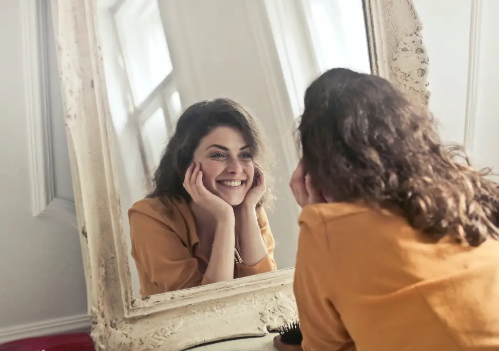 Pureday En kvinde med krøllet hår smiler varmt ved sit spejlbillede og fanger en følelse af naturlig velvære. Klædt i en gul skjorte hviler hun hagen på hænderne, mens blødt lys oplyser det rustikke indrammede spejl og forstærker rummets fredfyldte stemning.