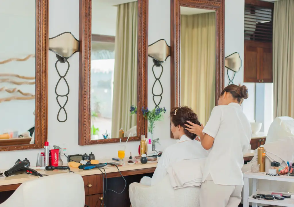 Pureday En frisør bliver set arrangere håret på en siddende klient i en salon. Rummet udstråler velvære med store spejle, elegante lysarmaturer og et udvalg af stylingværktøjer på disken, sammen med kamera, drink og forskellige produkter, der fremmer naturlig skønhed.